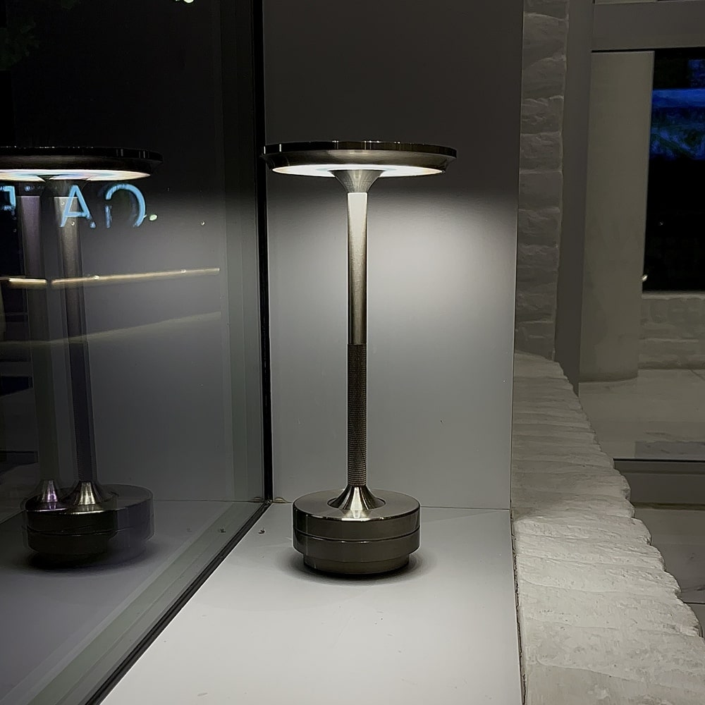 Silver cordless table lamp providing bright light, placed near a window, creating a reflective ambiance.