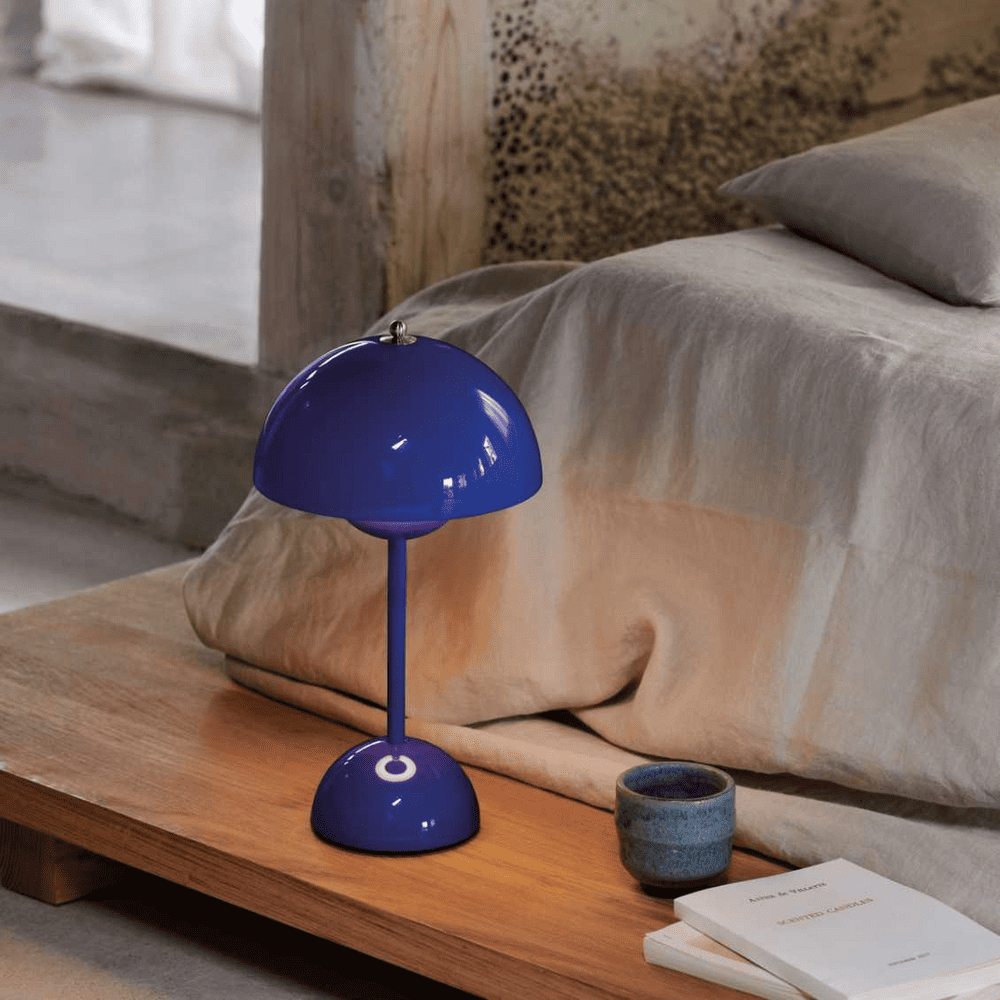 A vibrant blue modern table lamp placed on a wooden bedside table next to a ceramic mug and an open book. The setting creates a cozy, contemporary bedroom ambiance.