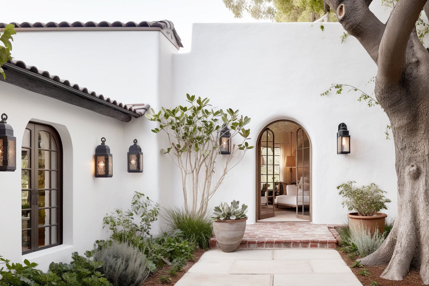 A Mediterranean-style home exterior featuring an arched doorway, white stucco walls, and black outdoor lanterns. The entrance is surrounded by lush greenery and a large tree, creating a welcoming and serene atmosphere.