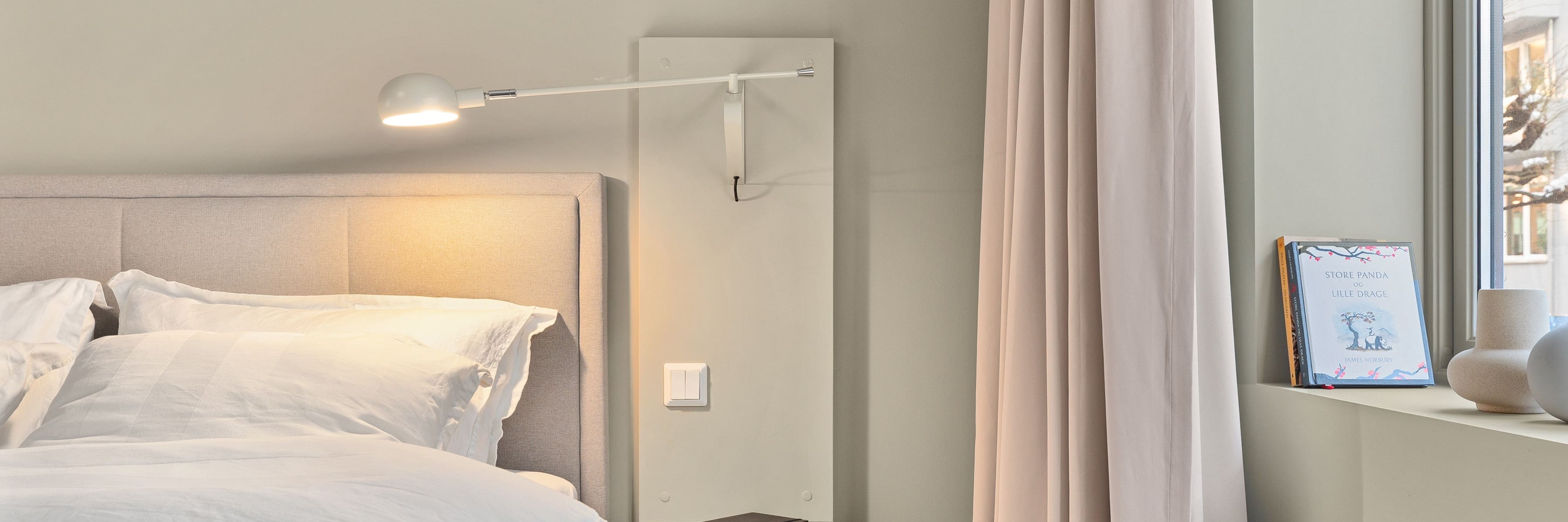 A minimalist bedroom with a beige upholstered headboard and wall-mounted reading light, a window with soft curtains, and a cozy atmosphere, ideal for relaxing and reading in bed.