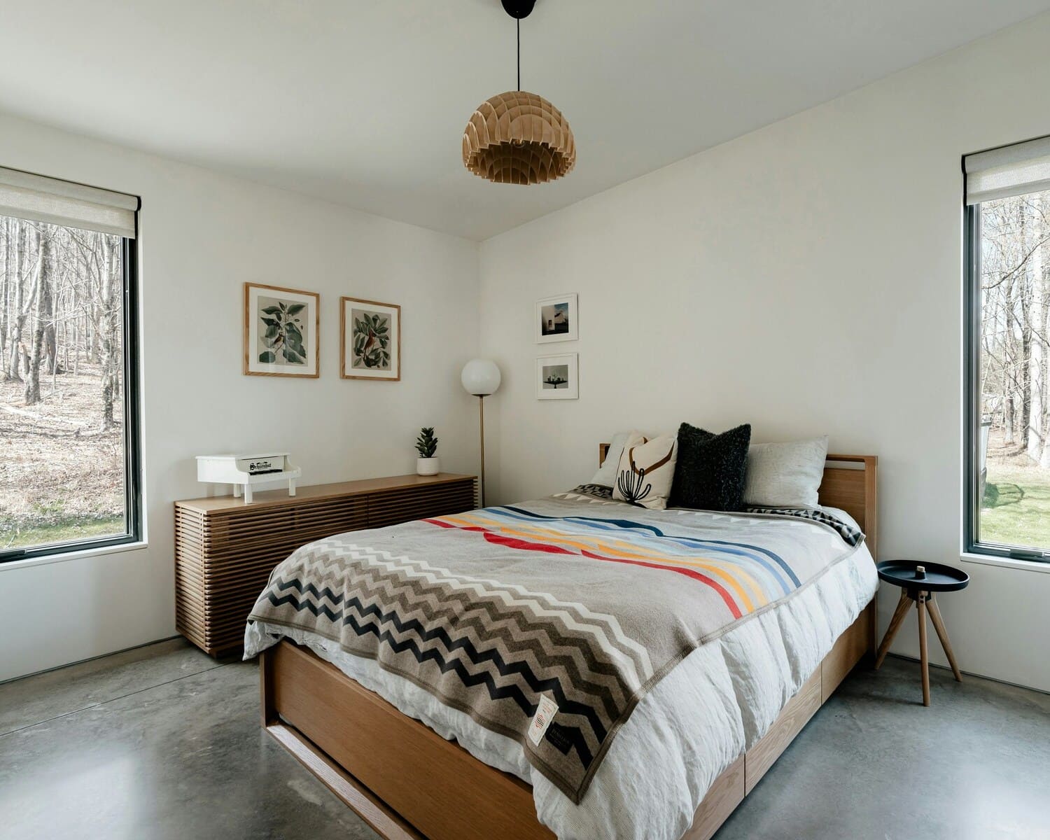 A minimalist Scandinavian bedroom featuring a wooden light pendant, simple decor, and natural lighting from large windows.