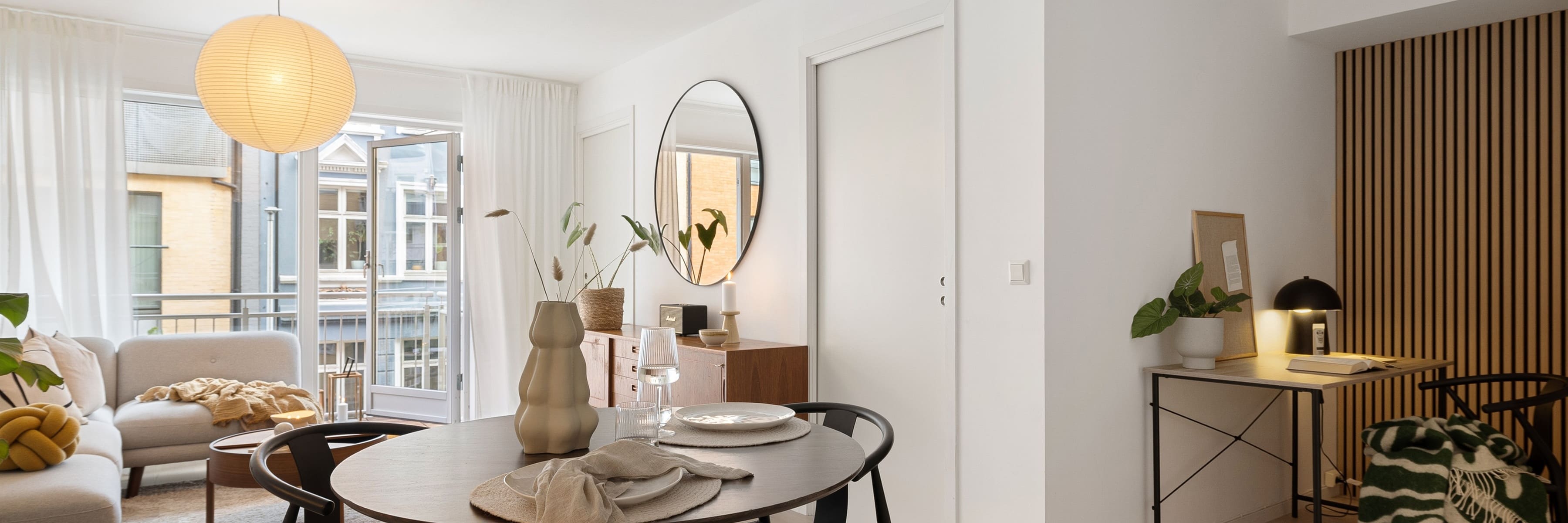 Scandinavian-style living room featuring a minimalistic round pendant light hanging above a dining table with black chairs and wooden cabinets. The space is bright with large windows and modern decor, including a round mirror and table decorations.