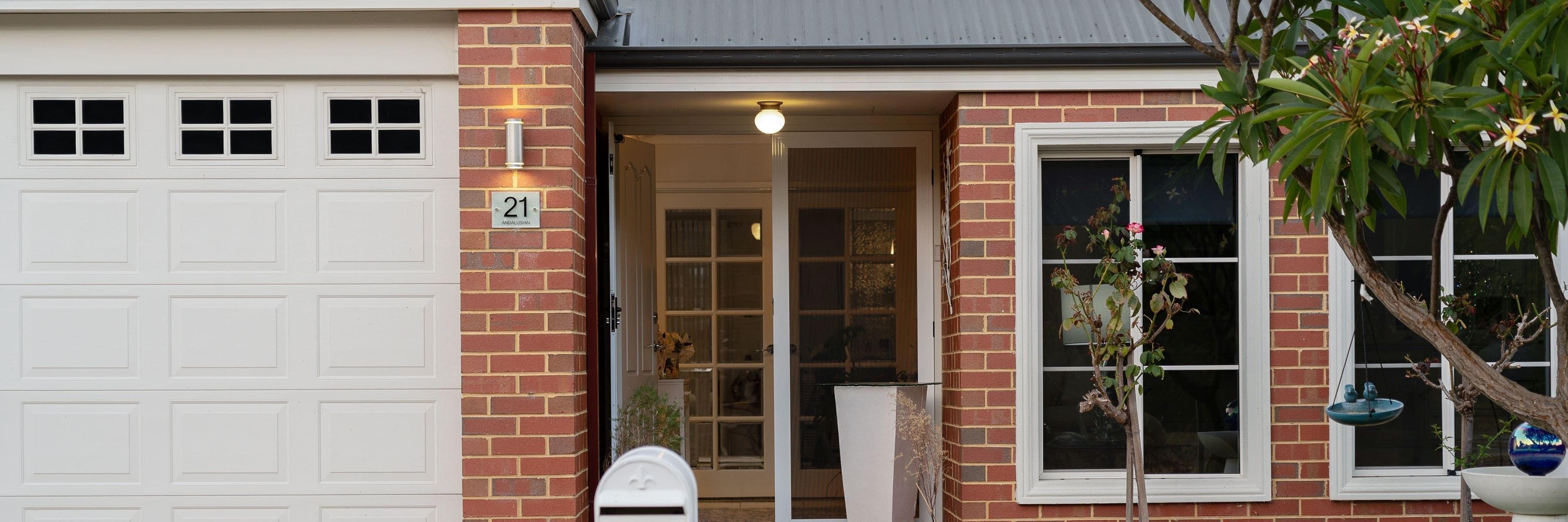 A modern home exterior with a red brick wall and white garage door, featuring a sleek outdoor wall light that illuminates the house entrance, enhancing security and curb appeal.