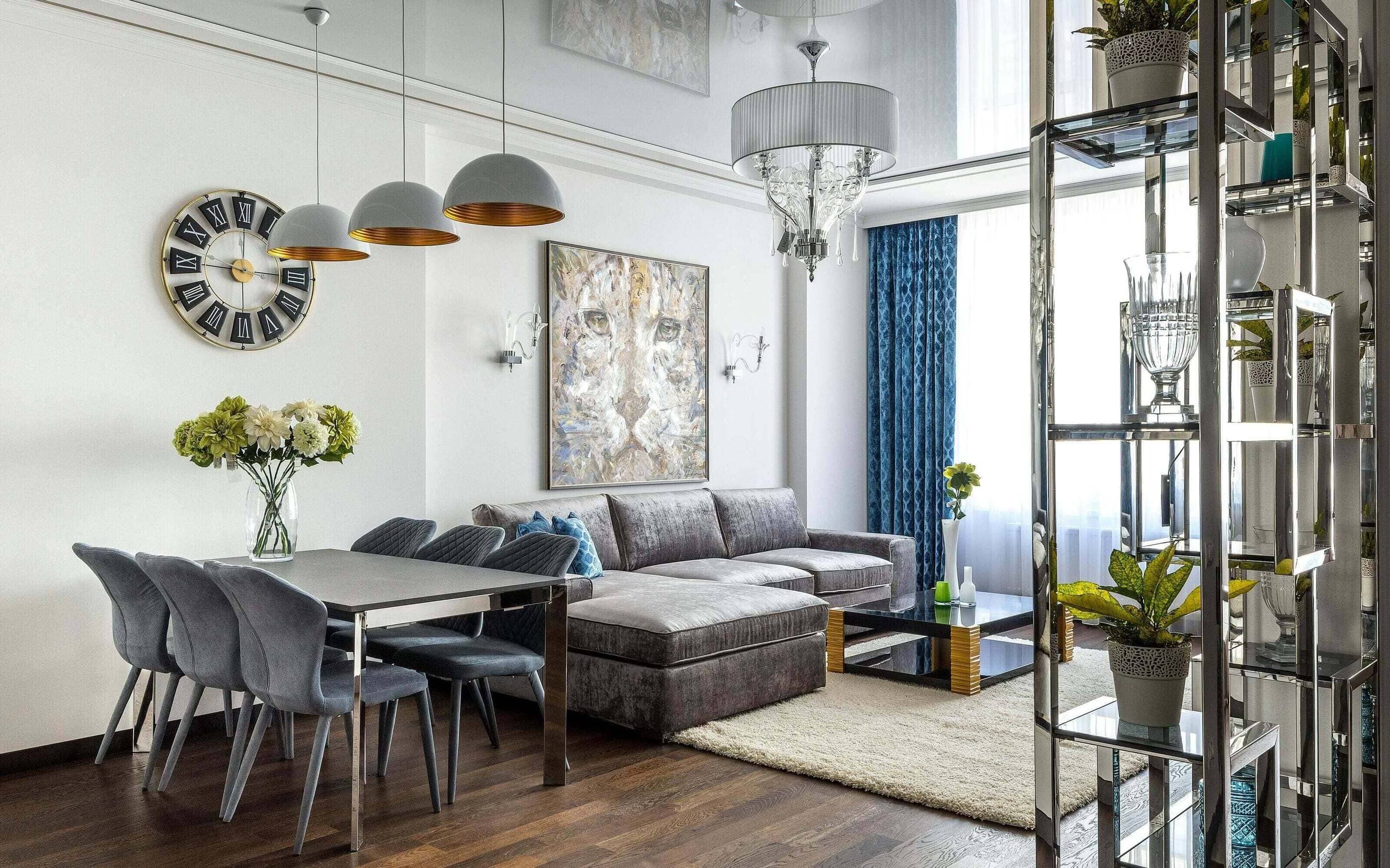 A modern living room interior featuring gray velvet sofa, a glass coffee table, pendant ceiling lights, and a dining area with gray upholstered chairs. The room is accented by a large wall clock and a lion-themed painting.