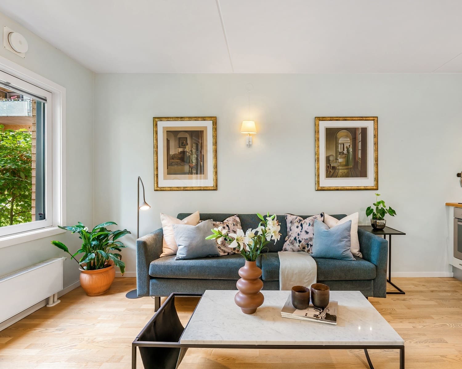 A modern living room with a teal sofa, decorated with patterned pillows, a wall sconce with a fabric shade, two framed art pieces, a floor lamp, and lush green plants, creating a fresh and inviting atmosphere.