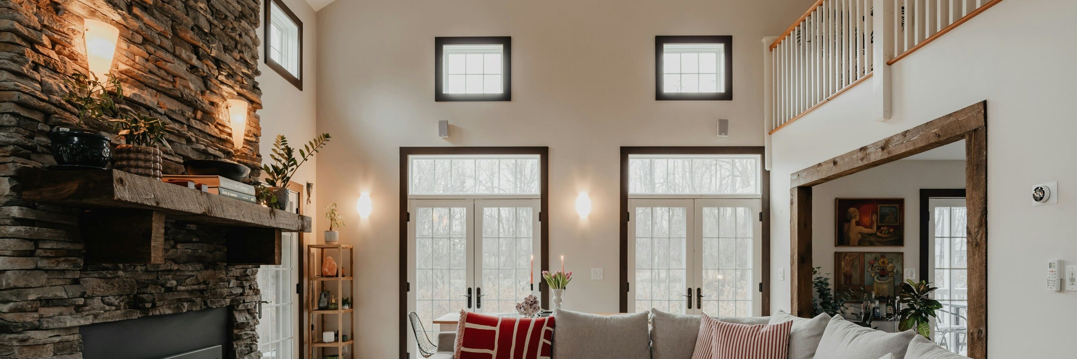 A rustic living room with high ceilings, stone fireplace, and wooden beams, featuring soft glowing wall sconces, a comfortable gray sofa with decorative pillows, and a cozy atmosphere.