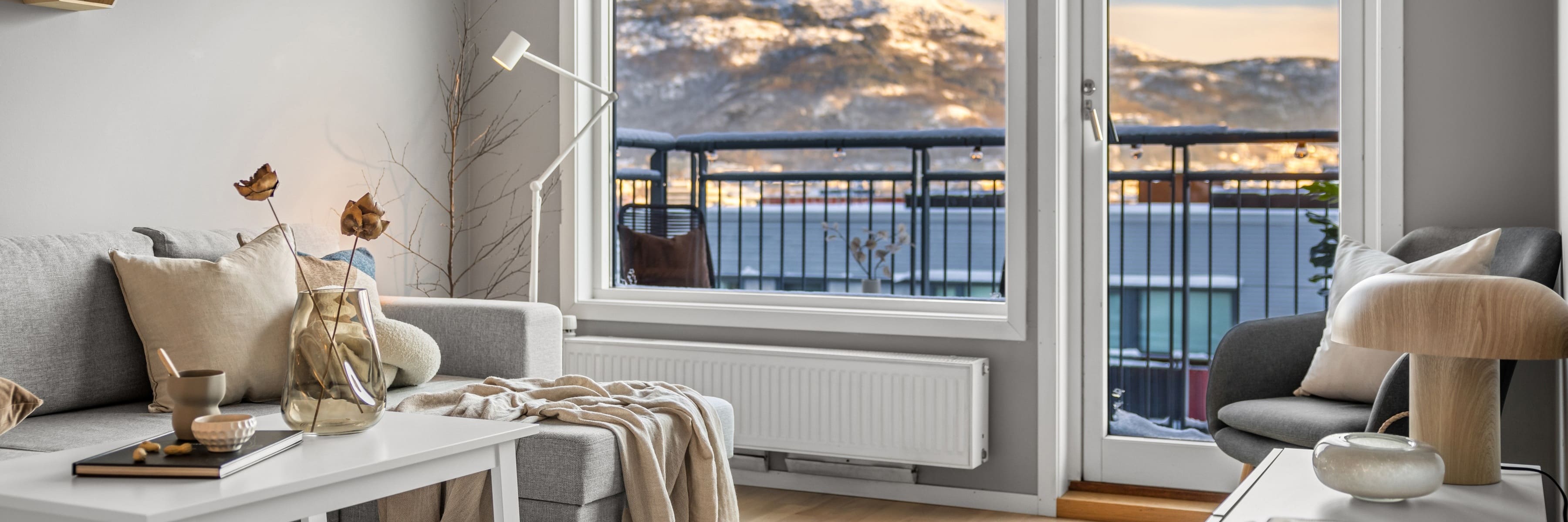 A Scandinavian-inspired living room with a neutral color palette, featuring a white floor lamp, a gray sofa, decorative pillows, a glass vase with dried flowers, and a view of a snowy landscape through large windows.